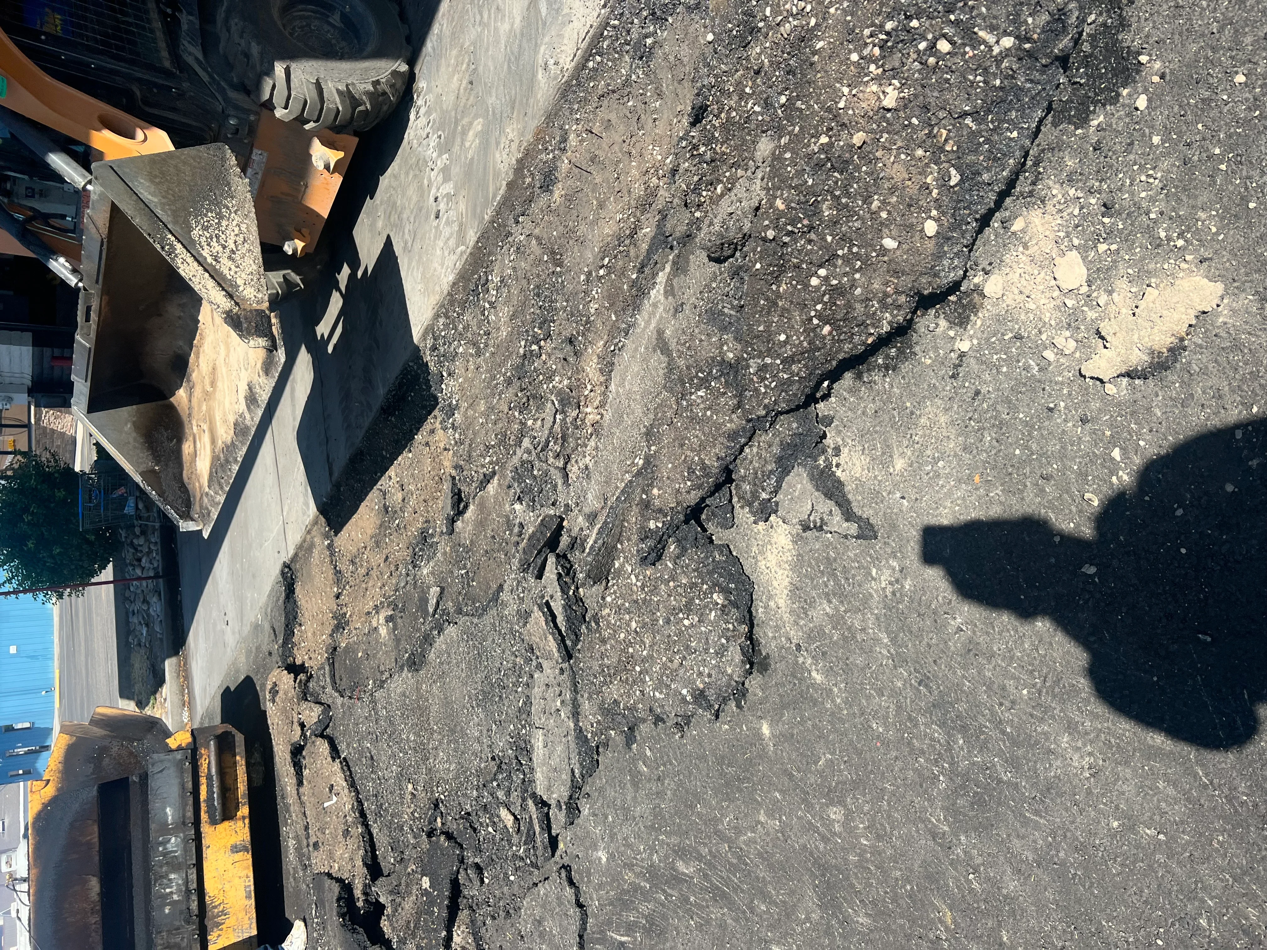 Excavator removing cracked and spalled asphalt pavement with visible base material exposed